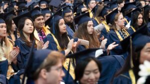 UC graduates seated in caps and gowns clapping.